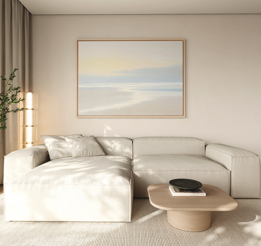 A minimalist living room featuring a light beige sectional sofa, wooden coffee table with black bowl, coastal accents like the Quiet Shores Canvas Art – Horizontal on the wall, light curtains, and a tall plant in the corner.