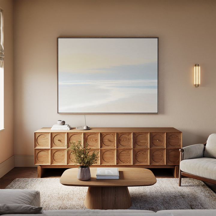 A living room with a beige wall, Quiet Shores Canvas Art – Horizontal above a wooden sideboard with circular patterns, modern coffee table, coastal decor accents like a floral vase, and a light gray sofa on a textured rug.