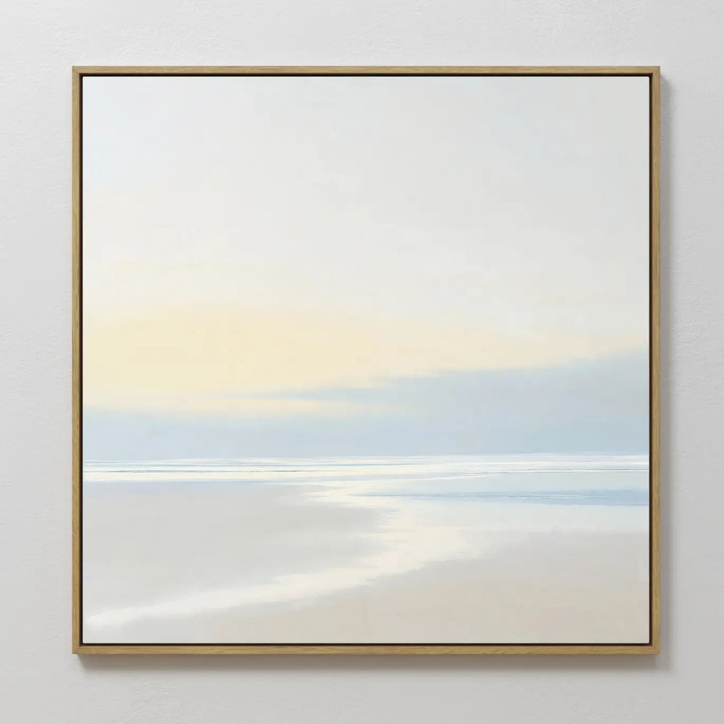 Quiet Shores Canvas Art features a minimalist seascape with soft pastels, a pale sky, and gentle clouds. Framed in thin gold and displayed on a white wall, its perfect coastal decor for any space.