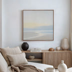 A minimal, modern living room features a beige sofa, neutral pillows, wooden sideboard, vases, books, and Quiet Shores Canvas Art on a white wall. Soft natural light enhances the cozy spaces coastal decor.