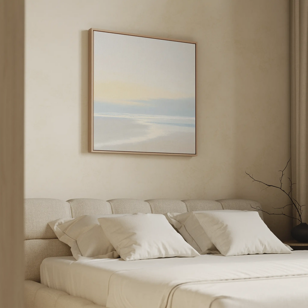 A minimal, modern bedroom features a beige upholstered bed with white bedding. Quiet Shores Canvas Art hangs above, enhancing the serene vibe, while soft natural light and beige curtains complement the tranquil coastal decor.
