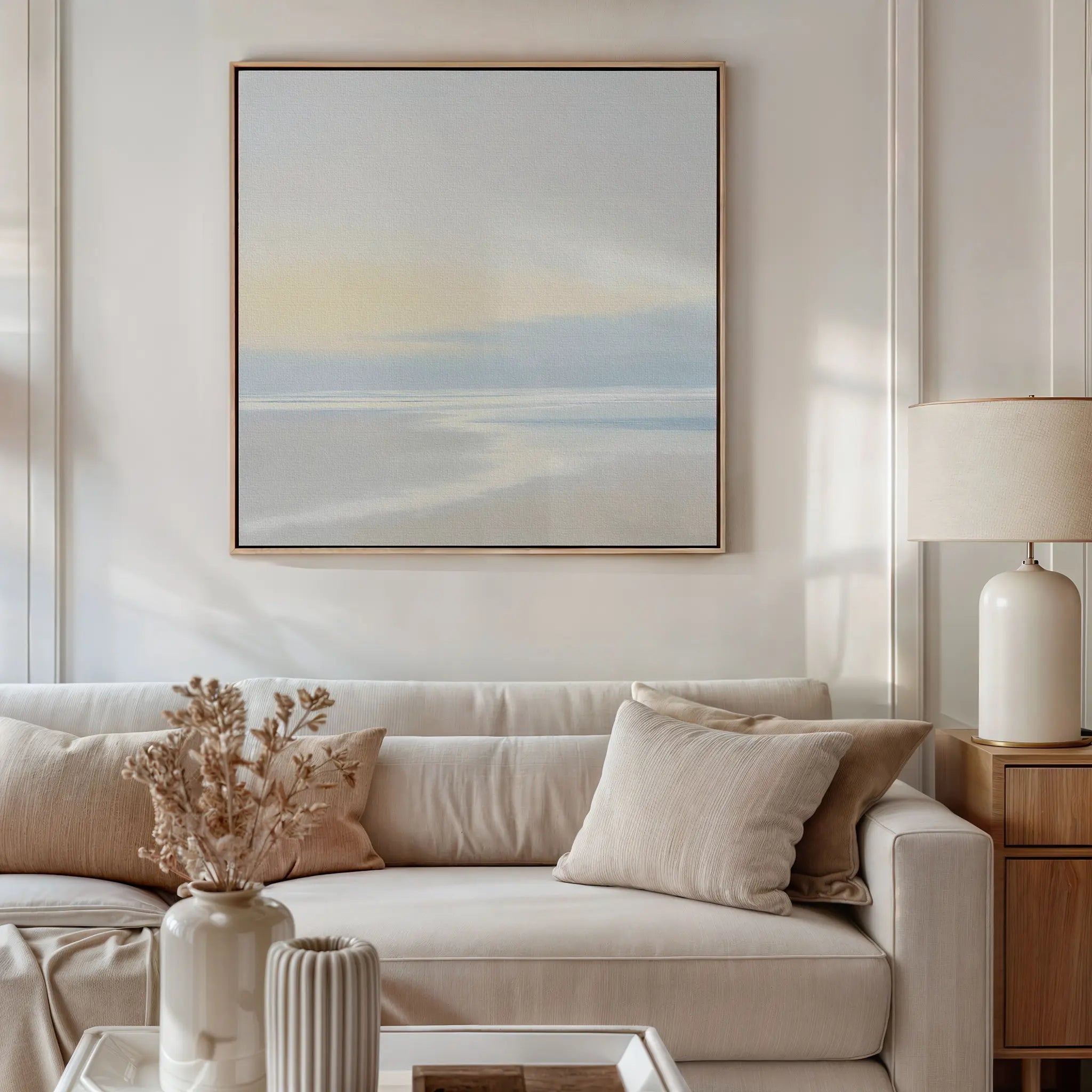 A modern living room features a neutral-toned sofa with beige cushions, wooden side table and lamp, vases of dried flowers on the coffee table, and Quiet Shores Canvas Art hanging above the seating area.
