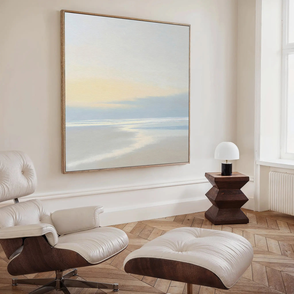 A modern, minimalist room features a white leather lounge chair with ottoman, a geometric side table with dome lamp, and the Quiet Shores Canvas Art above light wood herringbone floors, adding an elegant touch of coastal decor.