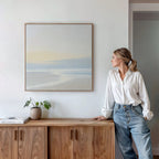 A woman in a white blouse and jeans leans against a wooden cabinet, admiring the Quiet Shores Canvas Art on the white wall. Nearby, a potted plant and books add to the serene coastal decor atmosphere.