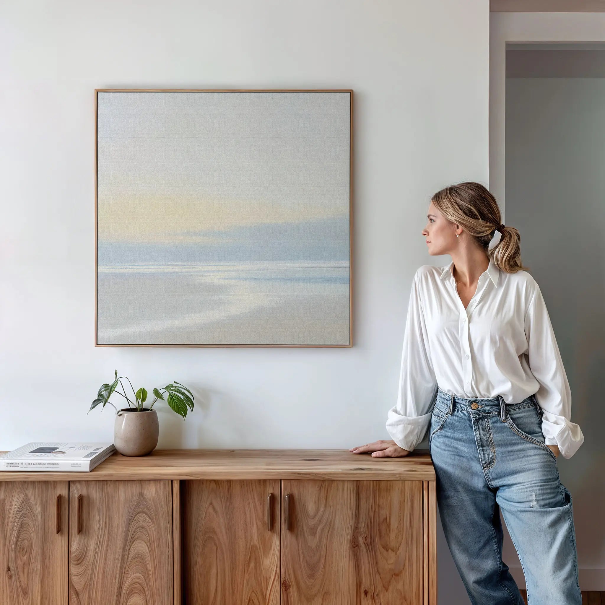 A woman in a white blouse and jeans leans against a wooden cabinet, admiring the Quiet Shores Canvas Art on the white wall. Nearby, a potted plant and books add to the serene coastal decor atmosphere.