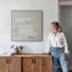 A woman in a white blouse and jeans leans against a wooden cabinet, admiring the Quiet Structure Canvas Art on the neutral wall. A small plant and books sit atop the cabinet in this minimal, modern room.