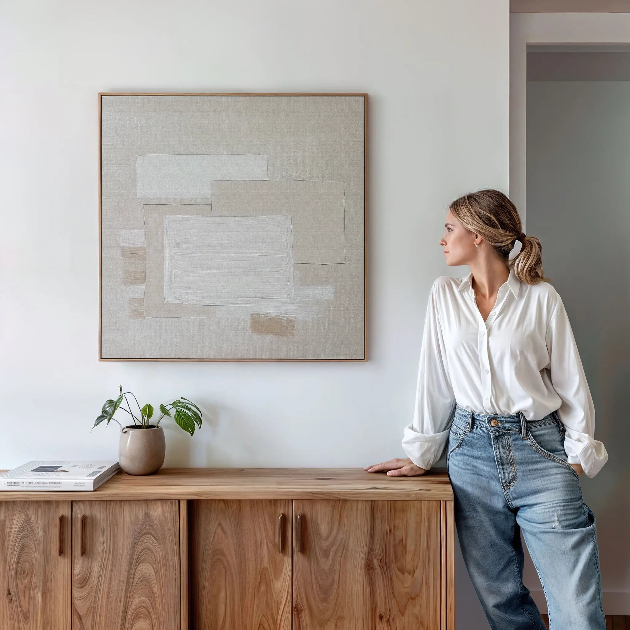 A woman in a white blouse and jeans leans against a wooden cabinet, admiring the Quiet Structure Canvas Art on the neutral wall. A small plant and books sit atop the cabinet in this minimal, modern room.