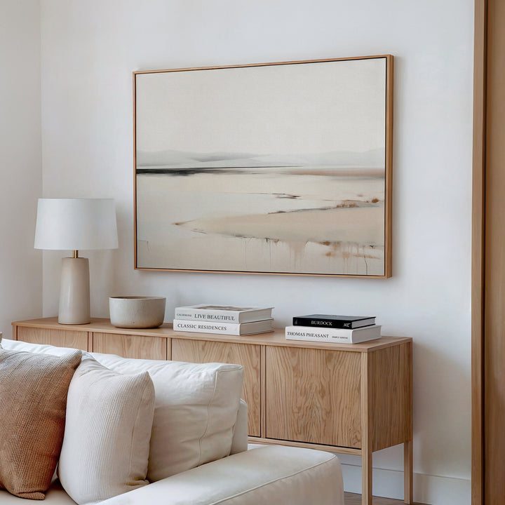 A modern living room with minimalist elegance features a light wood console table, white lamp, decor, stacked books, and the Quiet Valley Canvas Art – Horizontal hanging above a cream sofa with pillows.
