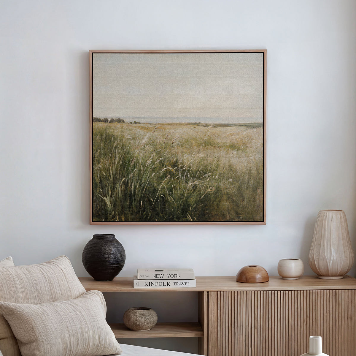 A serene living room with a beige sofa, vases, and books on a wooden console. Above, the Reeds by the River Canvas Art adds a minimalist touch with its depiction of calm grassy fields beneath a soft, cloudy sky.