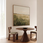 A minimalist dining area features a round wooden table, two cushioned chairs, and the Reeds by the River Canvas Art on a beige wall. Soft natural light through sheer curtains adds warmth to the living room decor.