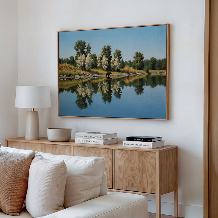 A modern living room with a light wood sideboard, white lamp, stacked books, and the Reflections of the Wild Canvas Art—featuring trees and a pond—hanging above a beige sofa for striking minimalist wall decor.