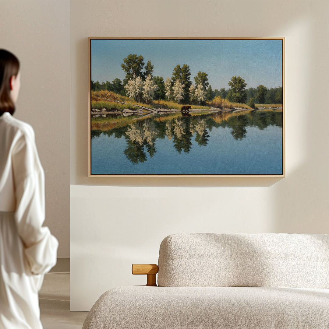 A person in a white robe stands in a bright, minimal room, admiring the Reflections of the Wild Canvas Art—large wall decor featuring trees mirrored on a calm lake—hanging above a beige sofa for an elegant minimalist touch.