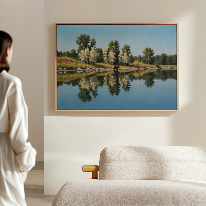 A person in a white robe stands in a bright, minimal room, admiring the Reflections of the Wild Canvas Art—large wall decor featuring trees mirrored on a calm lake—hanging above a beige sofa for an elegant minimalist touch.