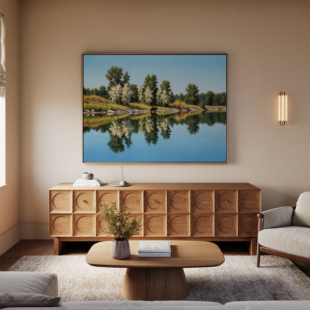 A cozy living room features a wooden coffee table, beige armchairs, a textured sideboard, and minimalist decor. The Reflections of the Wild Canvas Art landscape painting hangs on a neutral wall above a vase with flowers on the table.