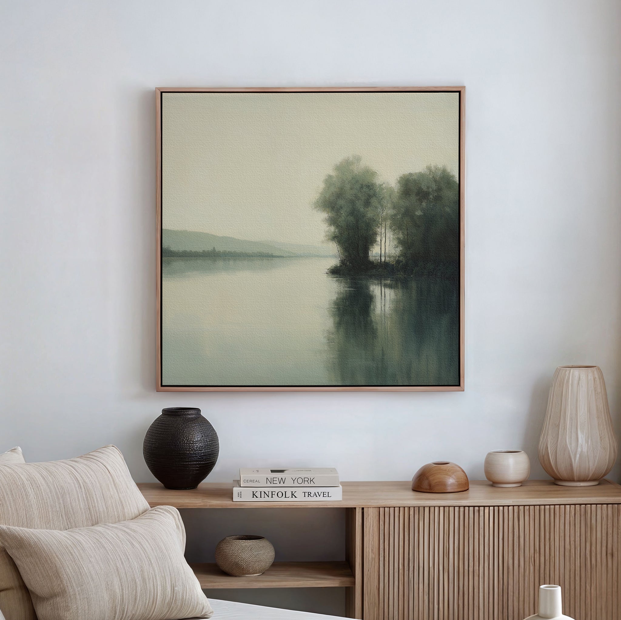 A serene living room with neutral decor features the Reflective River Canvas Art—vintage landscape of trees by a calm lake—displayed above a wooden sideboard adorned with vases, a lamp, and stacked books.