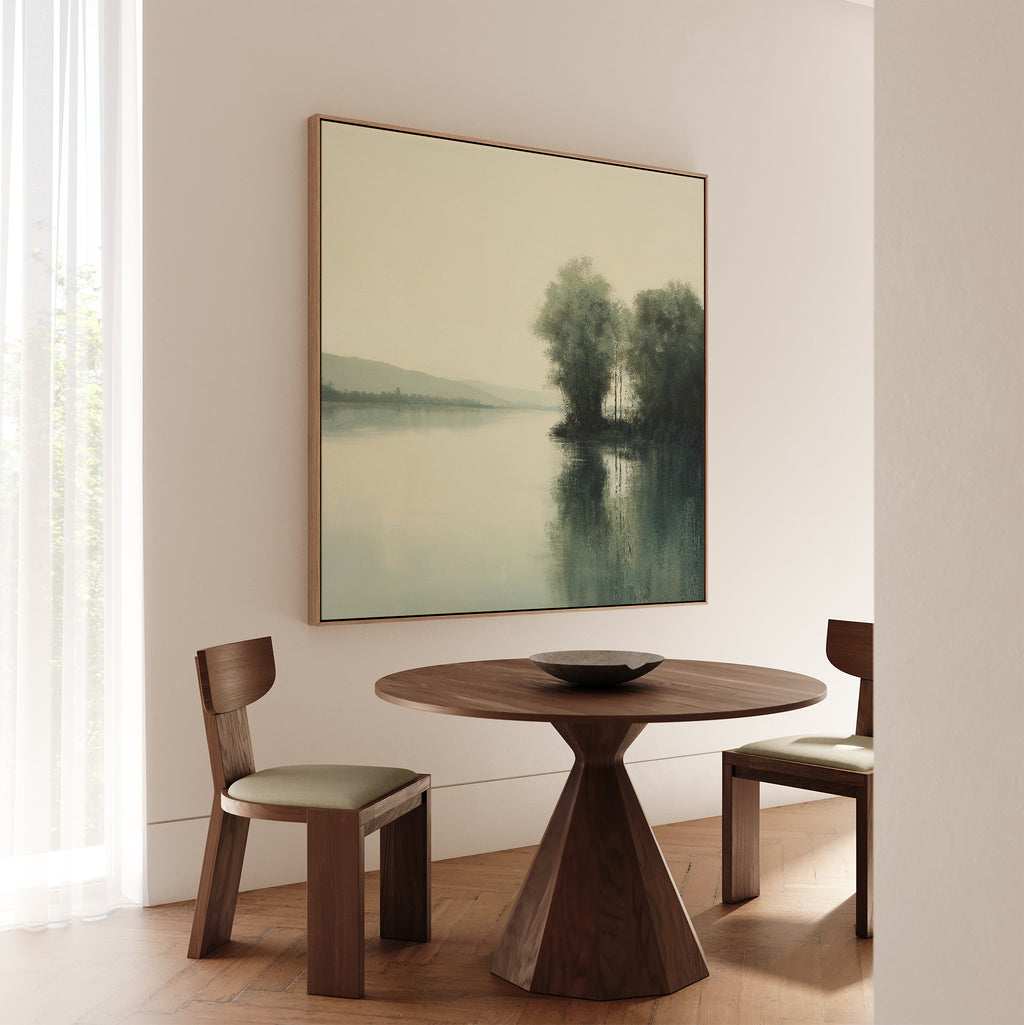 Minimalist dining area featuring a round wooden table, two cushioned wooden chairs, and the Reflective River Canvas Art—a serene wall piece depicting a lake with trees and reflections. Light streams in through sheer curtains.