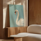The Regal Swan Canvas Art, featuring a white swan on blue, rests on a wooden console next to a white textured sofa and round table in a sunlit modern room with beige walls.