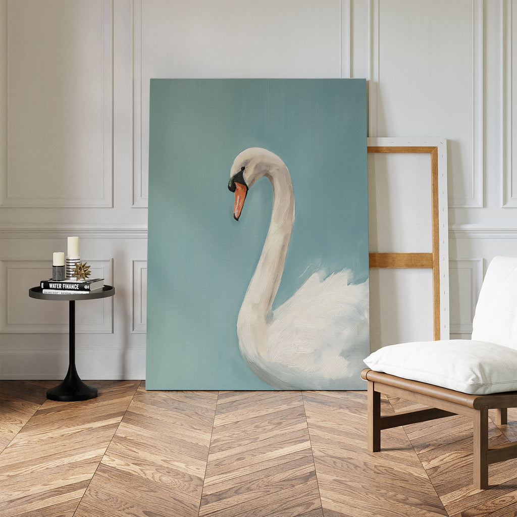 The Regal Swan Canvas Art, depicting a white swan against a blue background, leans on a white-paneled wall beside an empty frame, black side table with candles and books, and a cushioned chair on the wooden floor.