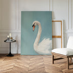 The Regal Swan Canvas Art, depicting a white swan against a blue background, leans on a white-paneled wall beside an empty frame, black side table with candles and books, and a cushioned chair on the wooden floor.