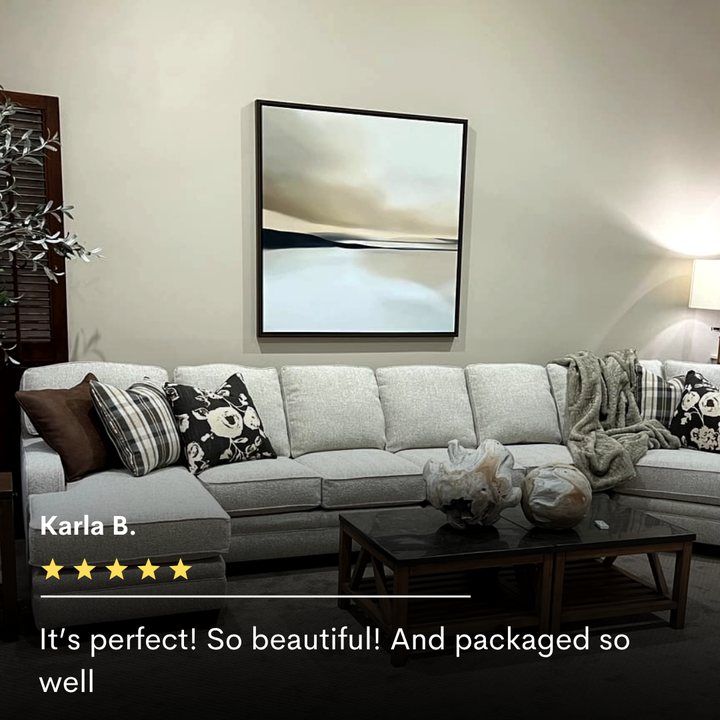 A modern living room with a light gray sectional, patterned pillows, cozy throw, glass coffee table with decor, soft lighting, and My Stores Desert Quietude Abstract Canvas Art. A 5-star review appears at the bottom.