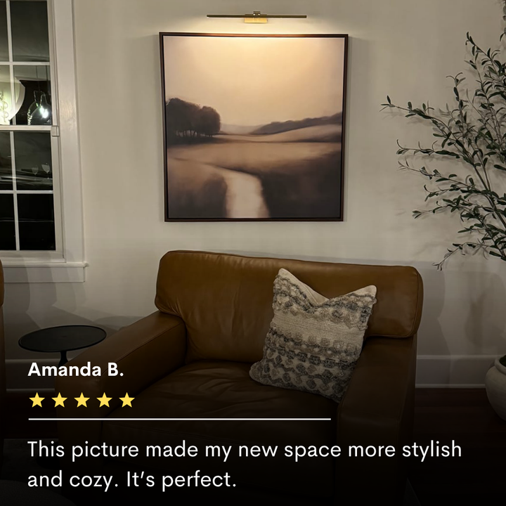 A brown leather chair with a textured pillow sits beneath Vintage Horizon Canvas Art—a framed landscape painting—on a light wall, lit by a picture light. A leafy plant is on the right, with a five-star review displayed below.