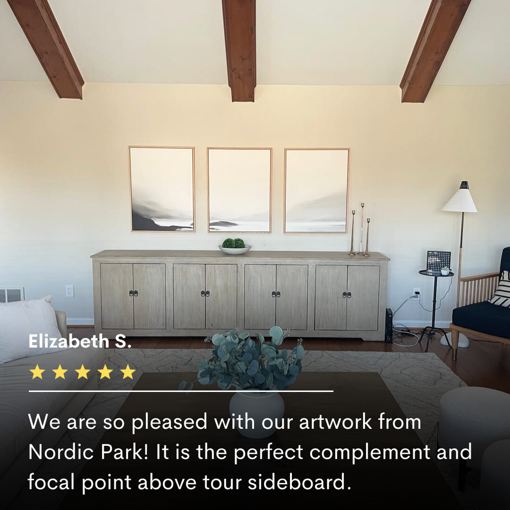 A light wood sideboard sits against a cream wall, topped with minimal decor. Above, the Serenity Peaks Set of 3 abstract landscape prints are evenly spaced. Exposed wooden beams and neutral modern furnishings create a serene balance.