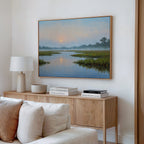 A cozy living room with a beige sofa, wooden sideboard, stacked books, a lamp, and large wall art—a framed Rising Stillness Canvas Art depicting a serene river scene at sunset—adds charm to the space.