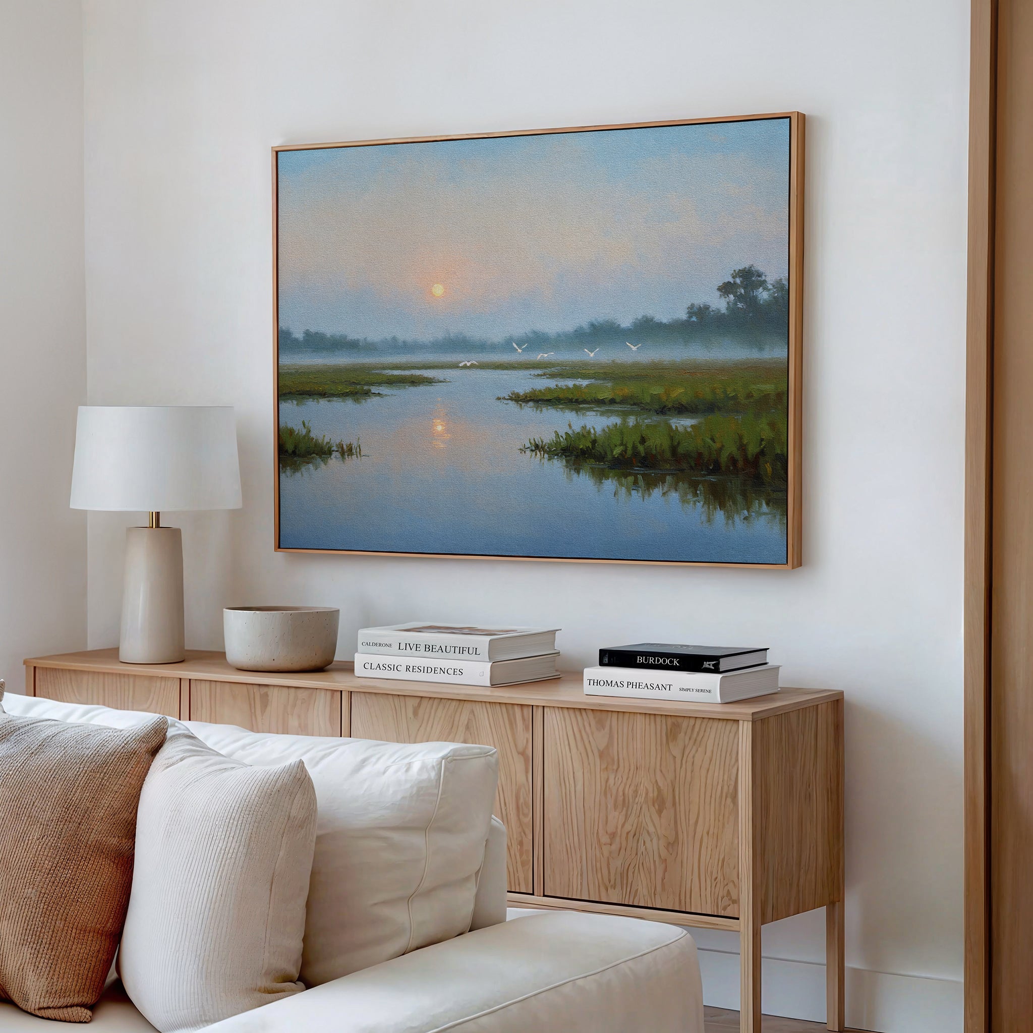 A cozy living room with a beige sofa, wooden sideboard, stacked books, a lamp, and large wall art—a framed Rising Stillness Canvas Art depicting a serene river scene at sunset—adds charm to the space.