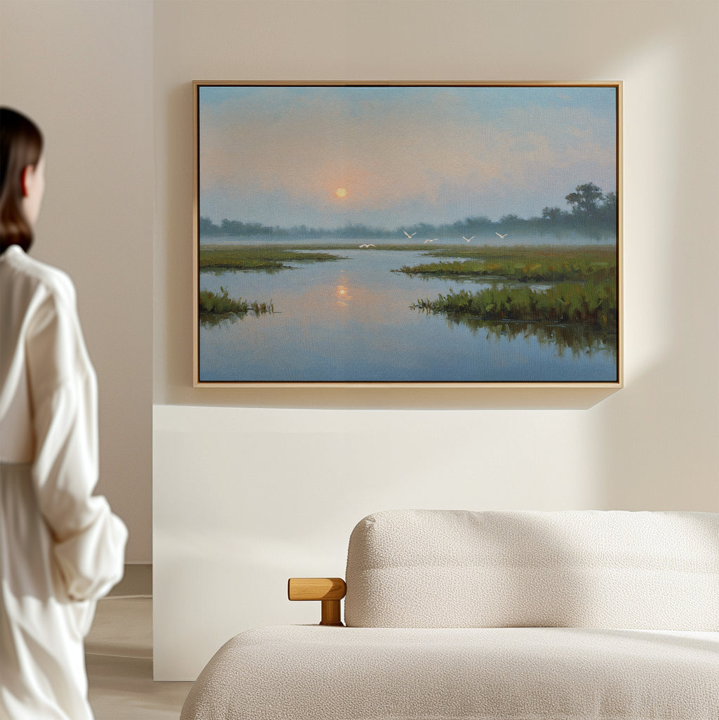 A person stands in a softly lit bedroom, gazing at the Rising Stillness Canvas Art—a serene river at sunset with birds and grassy banks. A beige sofa in the foreground adds calm elegance to the space.