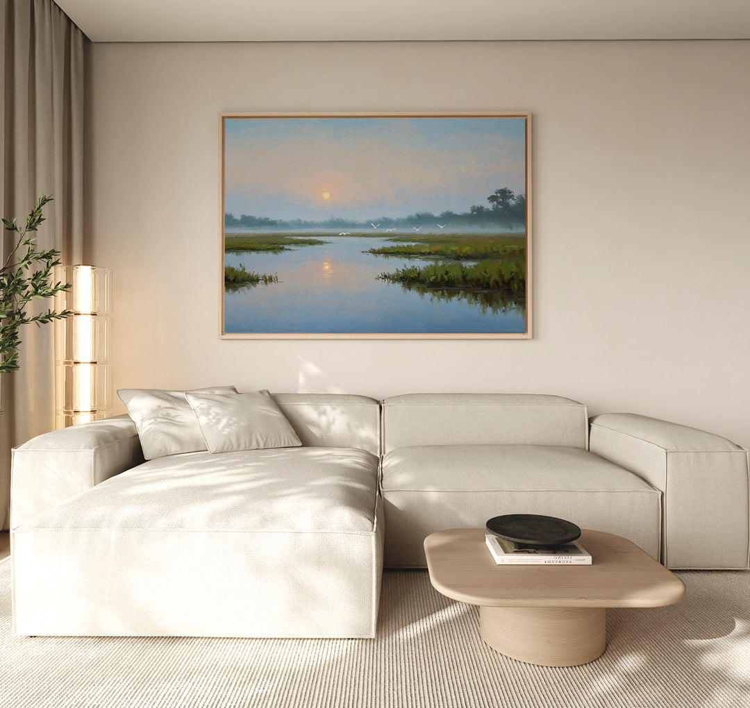 A minimalist living room with a cream-colored sectional sofa, a light wood coffee table, and the Rising Stillness Canvas Art—a striking sunrise over a river—hung above the sofa as a serene focal point.