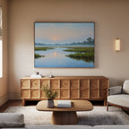 A cozy living room with a beige sofa, wooden coffee table, and textured sideboard. Above the sideboard hangs Rising Stillness Canvas Art depicting a serene river at sunset. A light fixture and books complete the inviting decor.