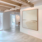 A minimalist room with white walls and light wooden beams features the Serene Expanse Abstract Canvas Art by My Store in neutral tones, while sunlight streams through windows and a dining area complements the modern living room decor.