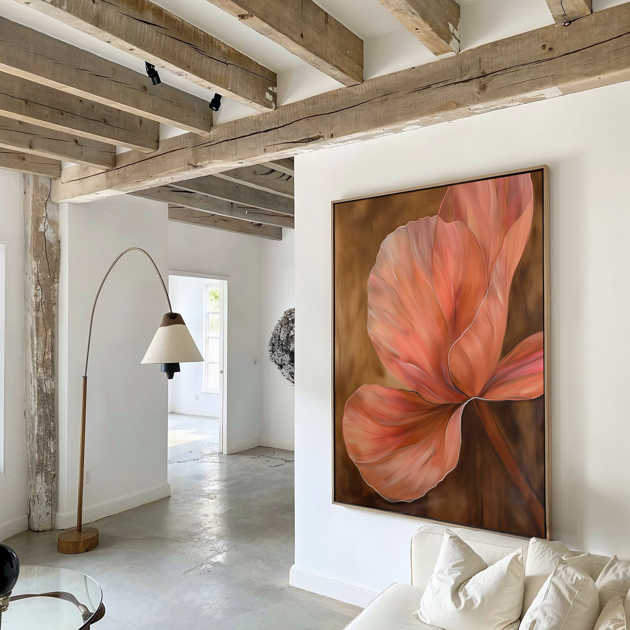 A modern living room with exposed wooden beams, white walls, and minimalist decor features the Blush Petals Abstract Canvas Art—a large painting of a pink flower on brown—hanging above a white couch. A curved floor lamp stands nearby.