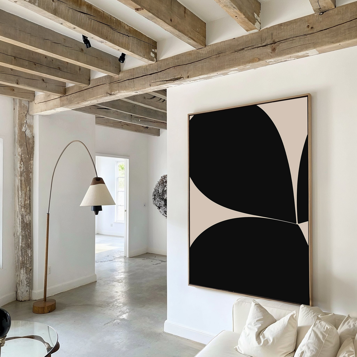 A modern living room with exposed wooden ceiling beams, white walls, contemporary decor including Black Botanical Abstract Canvas Art, a curved floor lamp, and a glimpse into a bright adjoining room.