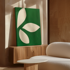 A modern living room features the Leaf Silhouette Abstract Canvas Art in green and white atop a sleek wooden cabinet, next to a textured white sofa and round table. Sunlight casts soft shadows, highlighting the space’s elegant simplicity.