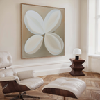 A modern living room features a cream lounge chair and ottoman, wooden herringbone flooring, a geometric side table with lamp, and Clover Harmony Abstract Canvas Art. Natural light pours in through a tall window, enhancing the decor.