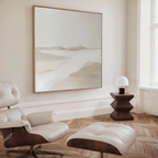 A minimalist living space featuring the Misty Vale Abstract Canvas Art—a striking large desert painting—paired with a white and wood lounge chair, geometric wooden side table, and a small white lamp by the window for effortless decor.