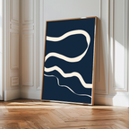 Blue Tranquility Abstract Canvas Art features wavy cream lines on a dark blue background, leaning against a white paneled wall in a sunny room with wooden parquet flooring—perfect for modern living room decor.
