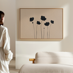 A woman in a white robe stands in a minimalistic living room, admiring the Botanic Abstract Canvas Art above a textured cream sofa. The artwork features black, abstract floral silhouettes on a light beige background.