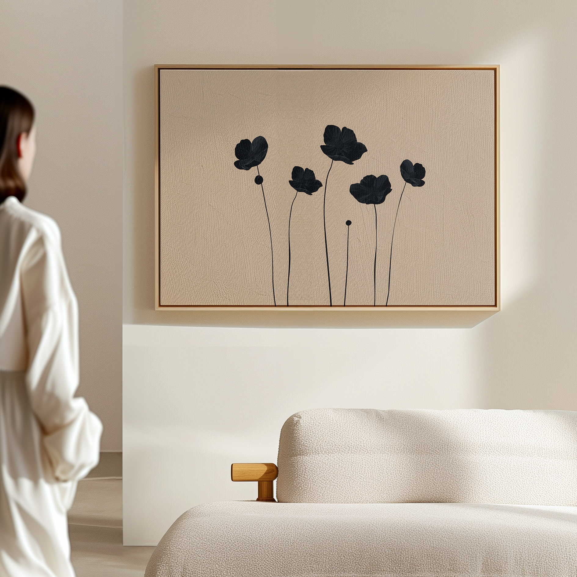 A woman in a white robe stands in a minimalistic living room, admiring the Botanic Abstract Canvas Art above a textured cream sofa. The artwork features black, abstract floral silhouettes on a light beige background.