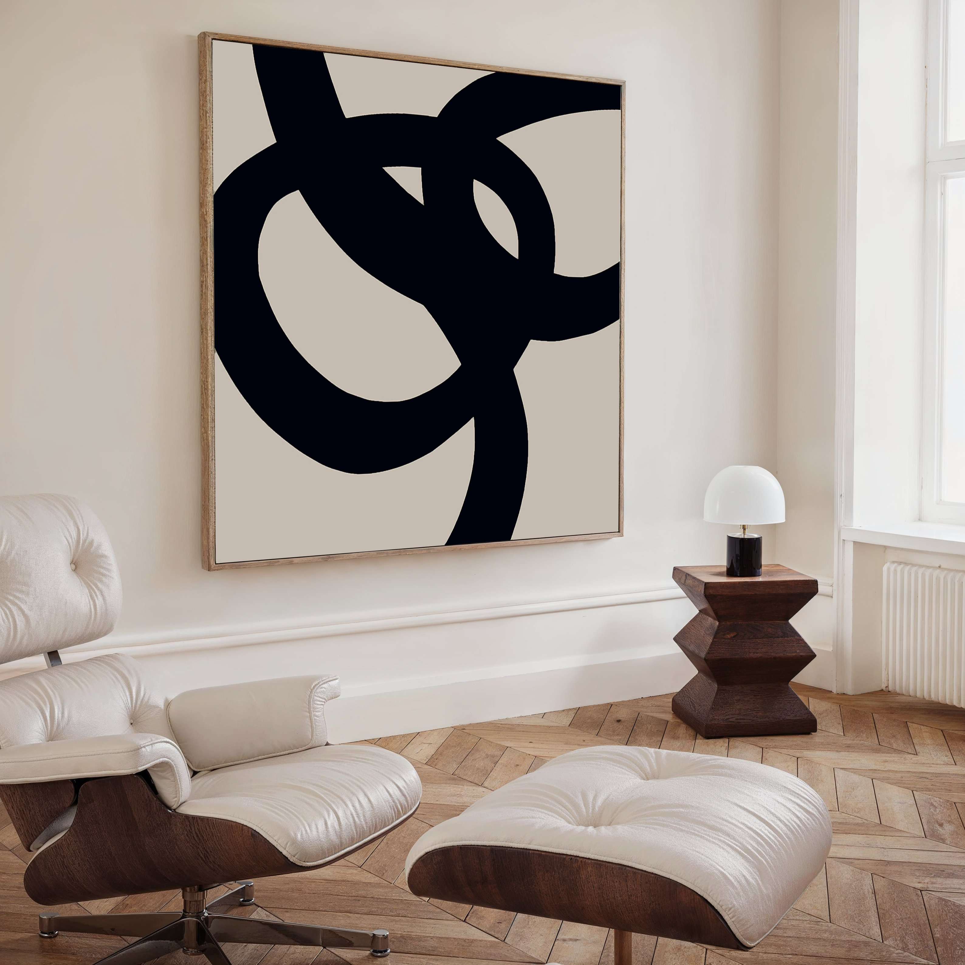 A modern, minimalist living room with a white lounge chair and ottoman, a geometric wooden side table with a white lamp, and Monochrome Stream Abstract Canvas Art featuring bold black loops on a beige background.