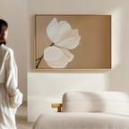 Dressed in light colors, a person stands in a minimalist room, admiring Tulips Abstract Canvas Art—a large framed piece with two white flowers on beige—displayed above a cream-colored sofa.