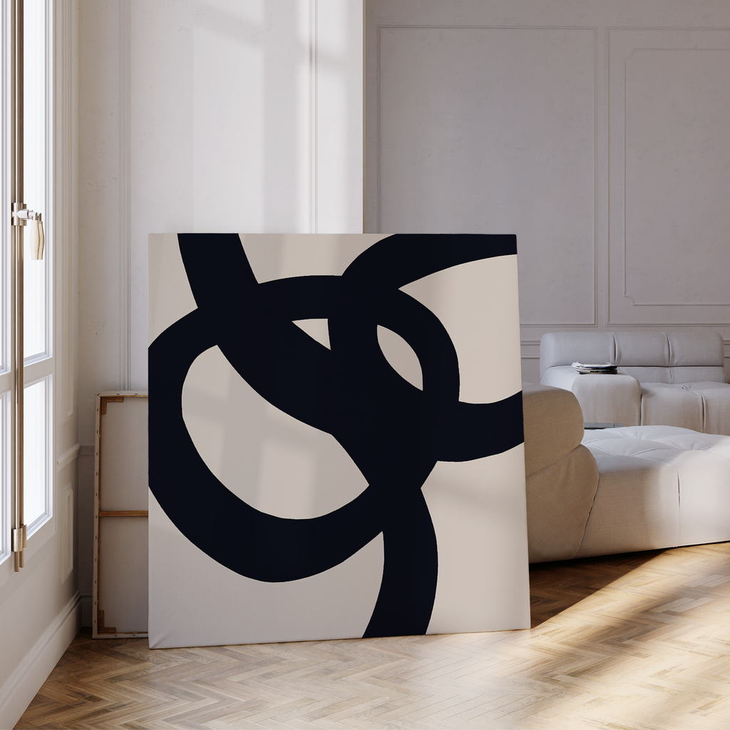 The Monochrome Stream Abstract Canvas Art, featuring bold black curves on white, rests on a wood floor in a bright living room, adding minimalist wall art beside a cream sofa and sunlight streaming through tall windows.