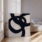The Monochrome Stream Abstract Canvas Art, featuring bold black curves on white, rests on a wood floor in a bright living room, adding minimalist wall art beside a cream sofa and sunlight streaming through tall windows.