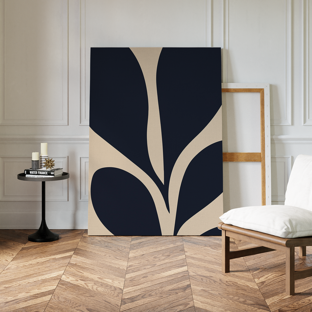 A minimalist room with the Botanical Ink Abstract Canvas Art—bold black shapes on beige—leaning against a white wall by an empty frame, a white armchair, and a table with candles and books; ideal for modern living room decor.