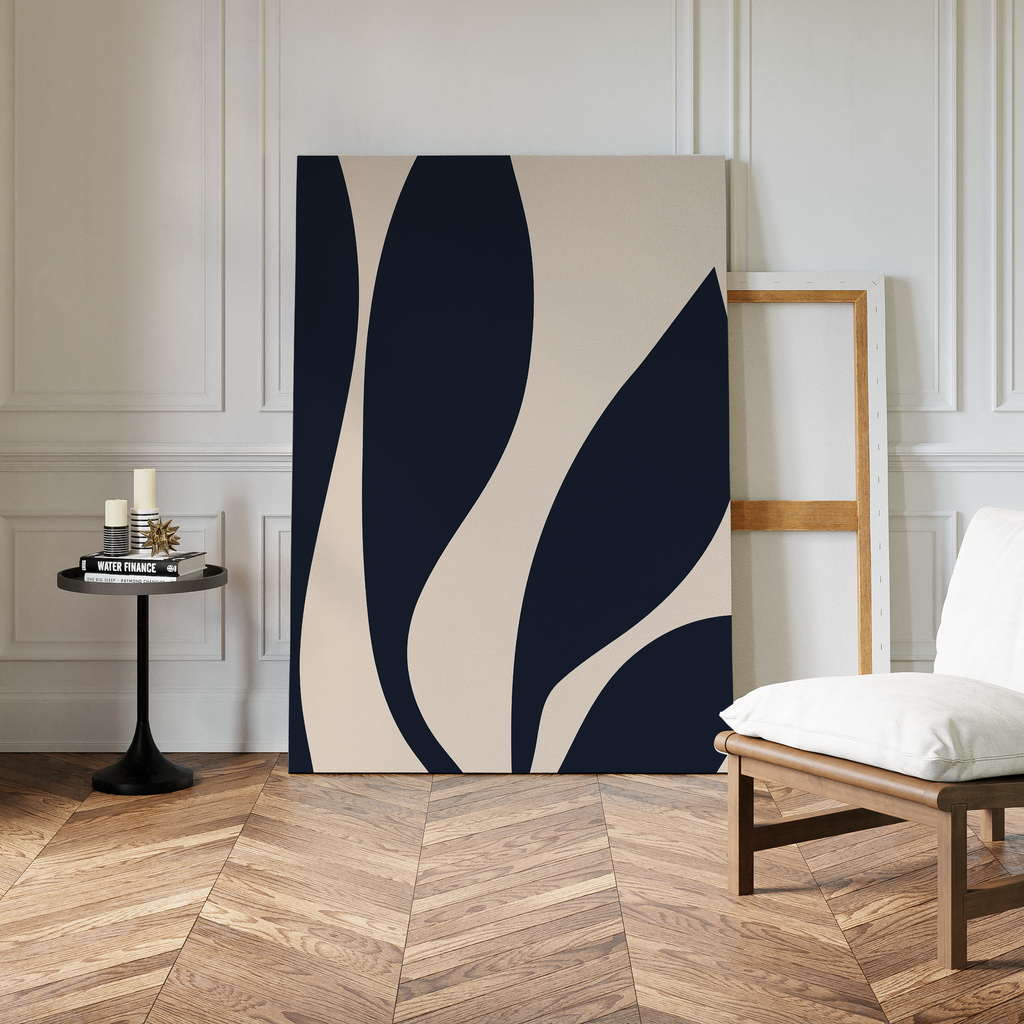 Dark Blue Leaves Abstract Canvas Art, featuring modern navy and beige curves, leans against a white paneled wall beside an empty frame, a cushioned white chair, and a round black table on a herringbone wood floor.