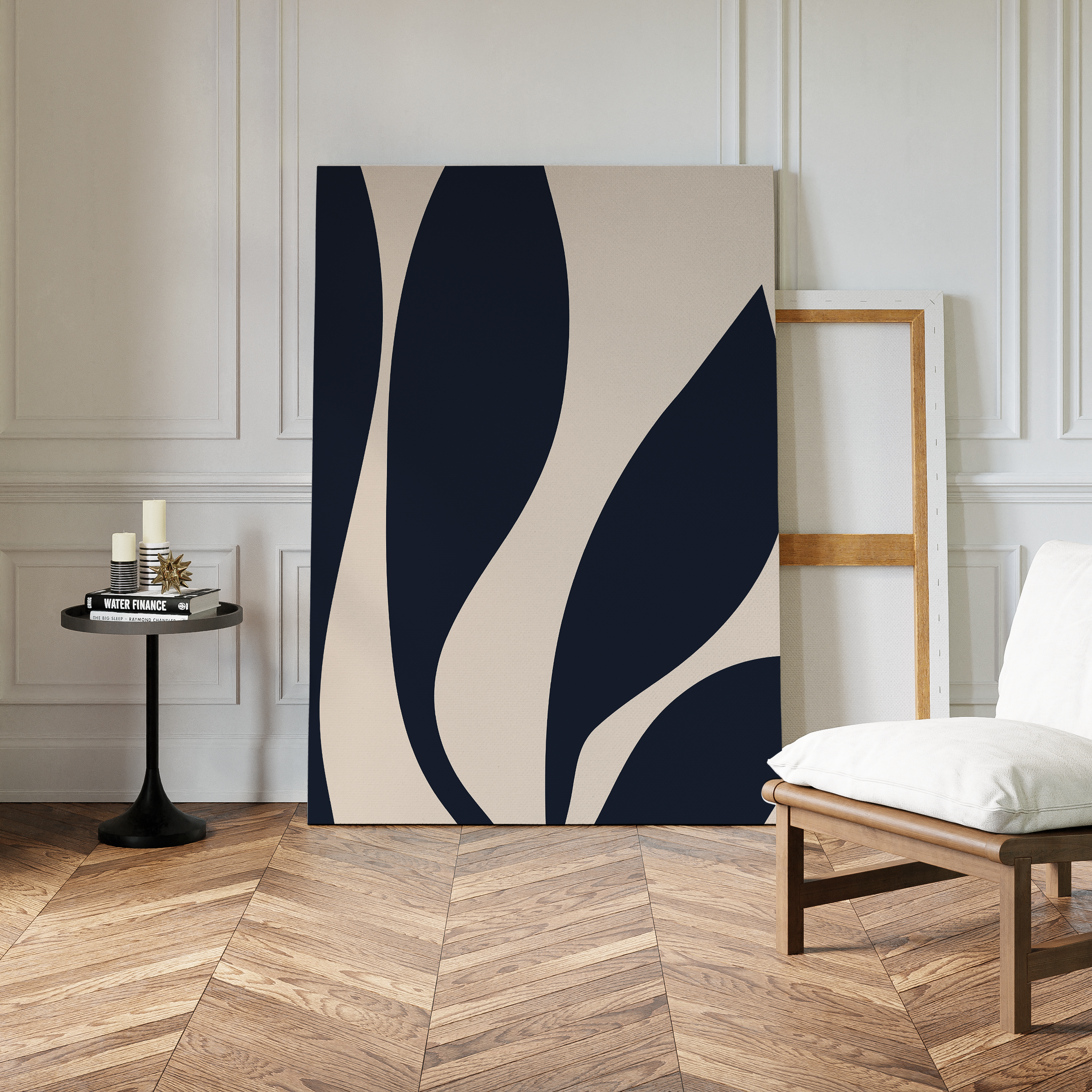 Dark Blue Leaves Abstract Canvas Art, featuring modern navy and beige curves, leans against a white paneled wall beside an empty frame, a cushioned white chair, and a round black table on a herringbone wood floor.