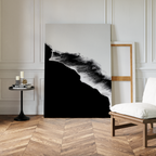 A modern room with chevron wood flooring features the Black Ocean Abstract Canvas Art propped against a wall in a wooden frame, alongside a white cushioned chair and a small black table with candles and books.