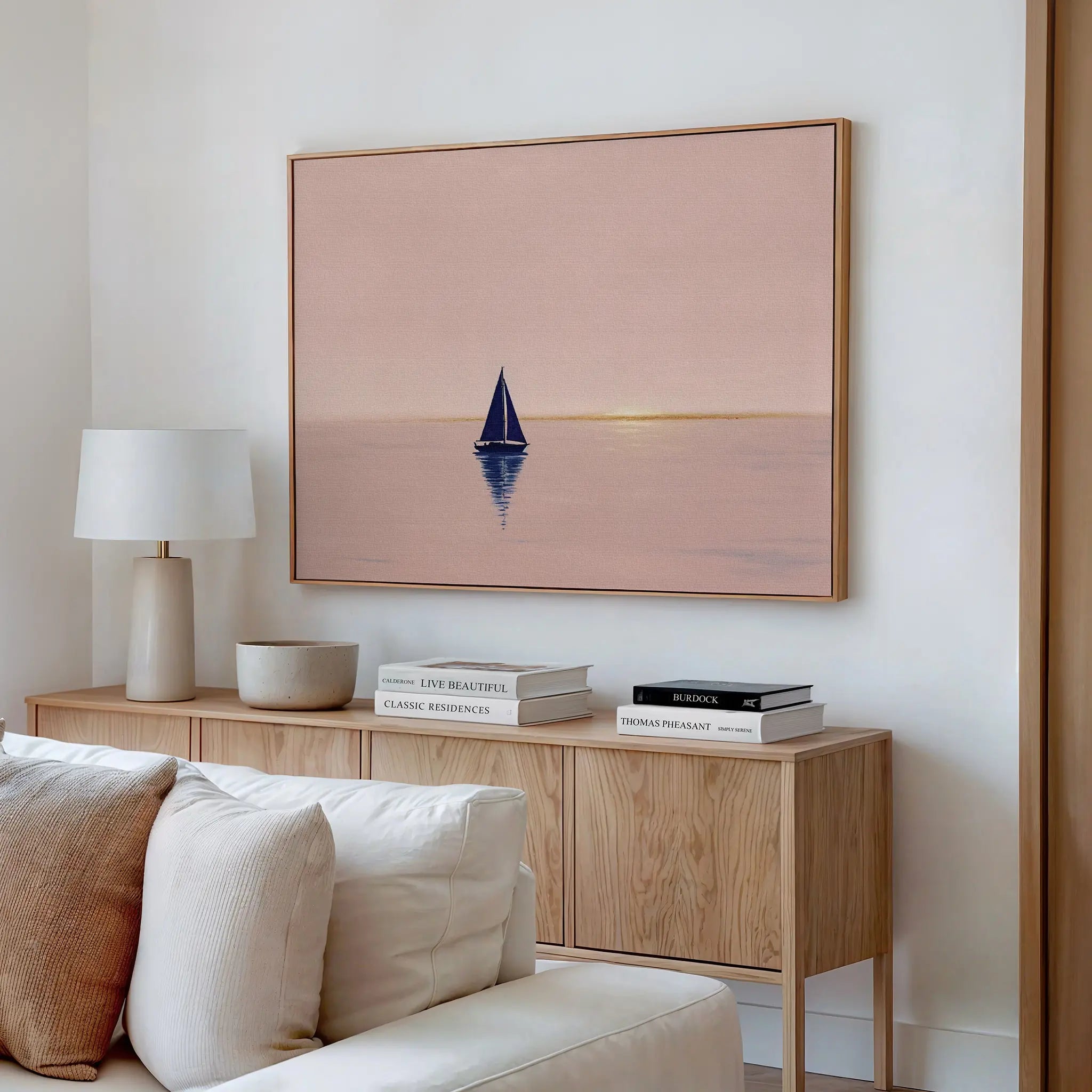 A minimalist living room with a light sofa and wooden sideboard features calming wall art—the Rose Harbor Canvas Art, a framed painting of a sailboat at sunset—elevating modern decor.