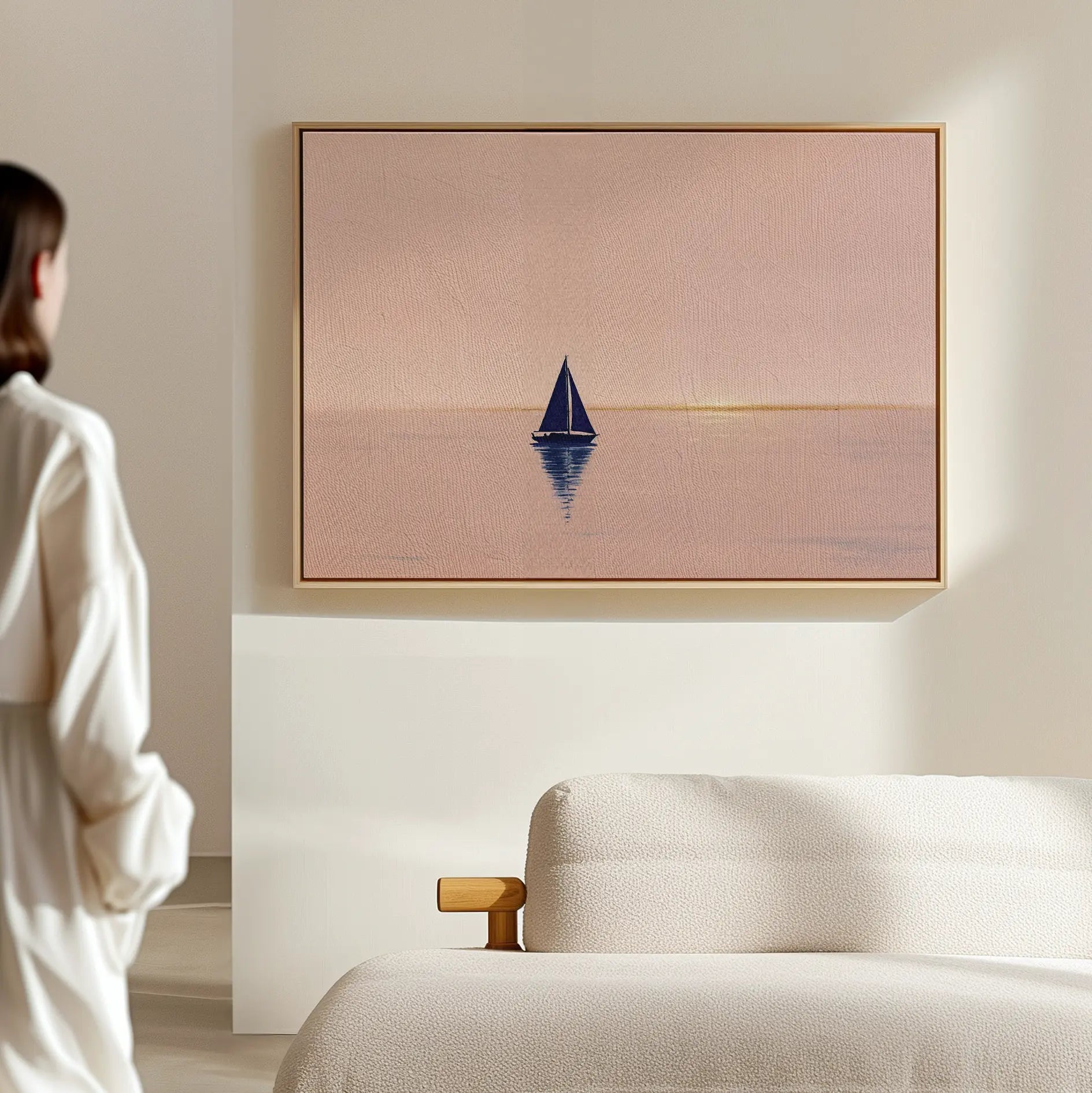 A woman stands in a modern room, admiring the Rose Harbor Canvas Art—a framed painting of a sailboat at sunset—displayed above a light-colored sofa.